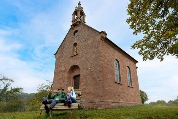 Wandernde legen die Beine auf einer Ruhebank vor einer Kapelle hoch und genie�en den Ausblick. Die Dreifaltigkeitskapelle �ber Reicholzheim auf dem LT 3 bildet den malerischen Hintergrund f�r eine wohlverdiente Pause. 