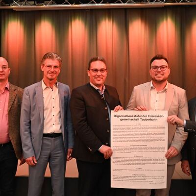 Bild vergr&ouml;&szlig;ern: Das Bild zeigt Landrat Christoph Schauder zusammen mit den neugewählten Sprechern, Bürgermeister Sebastian Mayer und Bürgermeister Michael Dieterich, sowie mit Bürgermeister Frank Menikheim und Bürgermeister Dr. Lukas Braun. Das beschlossene Organisationsstatut der neu gegründeten Interessengemeinschaft Tauberbahn wird als Poster gehoben.